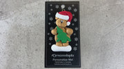 Tan bear in red festive hat holding green tree ornament, shown on backing card and Christmas tree.