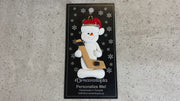 Ornament of white snowman in red hat holding a hockey stick and puck, shown on backing card and Christmas tree.