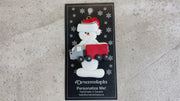 Ornament of white snowman in red hat holding a red semi truck, shown on backing card and Christmas tree.