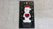 Ornament of white snowman in red hat holding a shiny red heart, shown on backing card and Christmas tree.