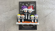 Family ornament with 6 black and white penguins in colourful festive hats, shown on backing card and Christmas tree.