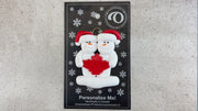 Family ornament with 2 white snowmen in red hats, gathered around a red maple leaf, shown on backing card and Christmas tree.