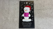 Ornament of white snowman in red hat holding a pink book, shown on backing card and Christmas tree.