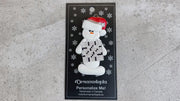 Ornament of white snowman in red hat holding large crossword puzzle, shown on backing card and Christmas tree.