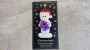 Ornament of white snowman in red hat holding a pink and purple birdhouse, shown on backing card and Christmas tree.