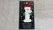 Ornament of white snowman in red hat holding green cheerleader pom-poms, shown on backing card and Christmas tree.