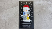 Ornament of white snowman in red hat holding a gardening glove and a shovel with flowers, shown on backing card and Christmas tree.