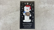 Ornament of white snowman in red hat holding dart board and one dart, shown on backing card and Christmas tree.