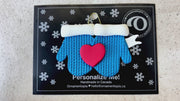A couple’s ornament of two mittens cradling a heart, shown on a backing card and Christmas tree.