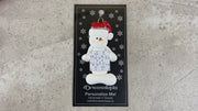 Ornament of white snowman in red hat holding a sudoku page, shown on backing card and Christmas tree.
