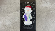 Ornament of white snowman in red hat holding a toothbrush and toothpaste, shown on backing card and Christmas tree.