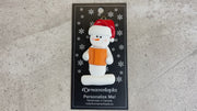Ornament of white snowman in red hat holding an orange book, shown on backing card and Christmas tree.