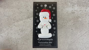Ornament of white snowman in red hat holding several playing cards, shown on backing card and Christmas tree.