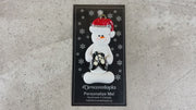 Ornament of white snowman in red hat holding black ballet shoes, shown on backing card and Christmas tree.