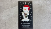 Ornament of white snowman in red hat holding a colourful guitar, shown on backing card and Christmas tree.
