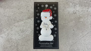 Ornament of white snowman in red hat with a round stomach that says “baby”, shown on backing card and Christmas tree.