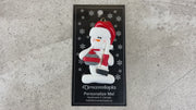 Ornament of white snowman in red hat holding red curling stone and broom, shown on backing card and Christmas tree.