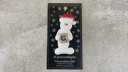 Ornament of white snowman in red hat holding a scrabble tile, shown on backing card and Christmas tree.
