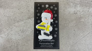Ornament of white snowman in red hat holding yellow curling stone and broom, shown on backing card and Christmas tree.