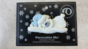 Ornament of white sleeping polar bear baby in a blue hat, shown on backing card and Christmas tree.