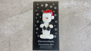 Ornament of white snowman in red hat carrying two dumbbells, shown on backing card and Christmas tree.