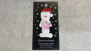 Ornament of white snowman in red hat holding a pink plastic crown, shown on backing card and Christmas tree.