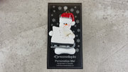 Ornament of white snowman in red hat holding a large white ice skate, shown on backing card and Christmas tree.