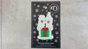 A couple’s ornament of two white snowmen holding a vibrant present, shown on backing card and Christmas tree.
