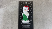 White snowman in red festive hat with green Christmas tree ornament, shown on backing card and Christmas tree.
