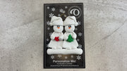 Family ornament with 2 white snowmen in hats, holding holiday objects, shown on backing card and decorated Christmas tree.