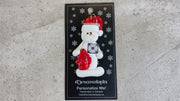 Ornament of white snowman in red hat holding a fire hydrant and firefighter badge, shown on backing card and Christmas tree.