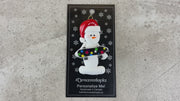 Ornament of white snowman in red hat holding a vibrant skateboard, shown on backing card and Christmas tree.