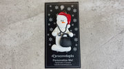 Ornament of white snowman in red hat holding a stethoscope and black medical bag, shown on backing card and Christmas tree.