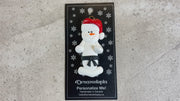 Ornament of white snowman in red hat wearing black belt, shown on backing card and Christmas tree.