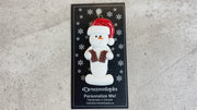 Ornament of white snowman in red hat holding two chocolate bars, shown on backing card and Christmas tree.