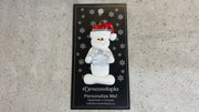 Ornament of white snowman in red hat holding clear plastic crown, shown on backing card and Christmas tree.