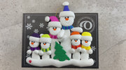 Family ornament with 6 white snowmen in bright scarves, with green Christmas tree, shown on backing card and Christmas tree.