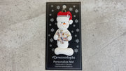 Ornament of white snowman in red hat holding a puzzle piece, shown on backing card and Christmas tree.