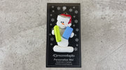 Ornament of white snowman in red hat holding vibrant yoga mat and block, shown on backing card and Christmas tree.