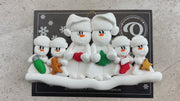 Family ornament with 2 large and 4 small snowmen in hats, holding holiday objects, shown on backing card and Christmas tree.