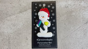 Ornament of white snowman in red hat holding a pickleball paddle and yellow pickleball, shown on backing card and Christmas tree.