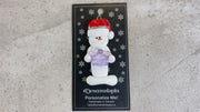 Ornament of white snowman in red hat holding purple plastic crown, shown on backing card and Christmas tree.