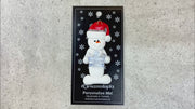 Ornament of white snowman in red hat holding a blue plastic crown, shown on backing card and Christmas tree.