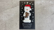 Ornament of white snowman in red hat holding cowboy boots, shown on backing card and Christmas tree.