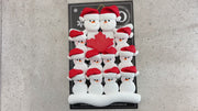 Family ornament with 12 white snowmen in red hats, gathered around a red maple leaf, shown on backing card and Christmas tree.