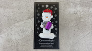 Ornament of white snowman in red hat holding a spool of thread and scissors, shown on backing card and Christmas tree.