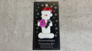 Ornament of white snowman in red hat holding a microphone and pink music book, shown on backing card and Christmas tree.