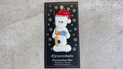 Ornament of white snowman in red hat holding a fishing pole and colourful fish, shown on backing card and Christmas tree.