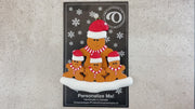 Family ornament with 1 large and 3 small brown gingerbread figures in red Christmas hats, shown on backing card and Christmas tree.
