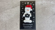 Ornament of white snowman in red hat holding a black camera, shown on backing card and Christmas tree.